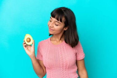 Young mixed race woman holding an avocado isolated on blue background looking to the side and smiling