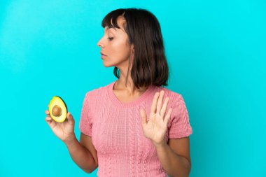Young mixed race woman holding an avocado isolated on blue background making stop gesture and disappointed