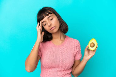 Young mixed race woman holding an avocado isolated on blue background with headache
