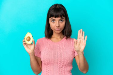 Young mixed race woman holding an avocado isolated on blue background making stop gesture