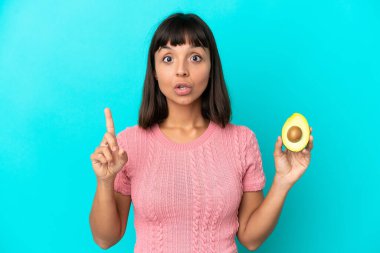 Young mixed race woman holding an avocado isolated on blue background intending to realizes the solution while lifting a finger up