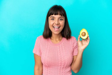 Young mixed race woman holding an avocado isolated on blue background with surprise facial expression