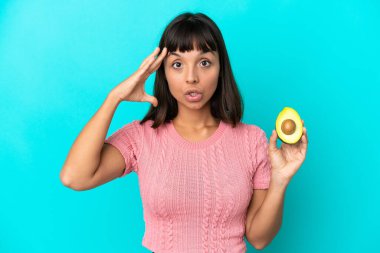 Young mixed race woman holding an avocado isolated on blue background with surprise expression