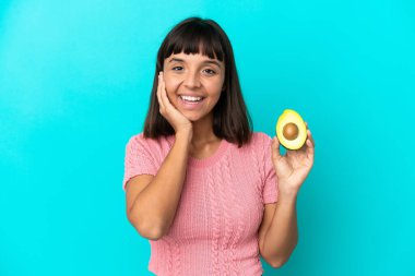 Young mixed race woman holding an avocado isolated on blue background with surprise and shocked facial expression