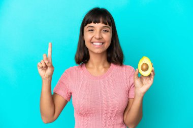 Young mixed race woman holding an avocado isolated on blue background showing and lifting a finger in sign of the best