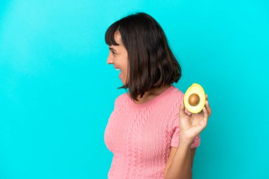 Young mixed race woman holding an avocado isolated on blue background laughing in lateral position