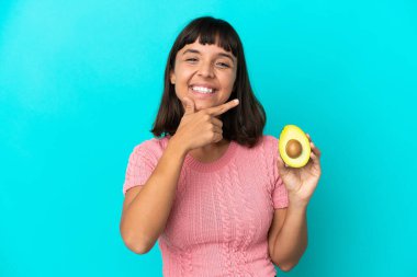 Young mixed race woman holding an avocado isolated on blue background happy and smiling