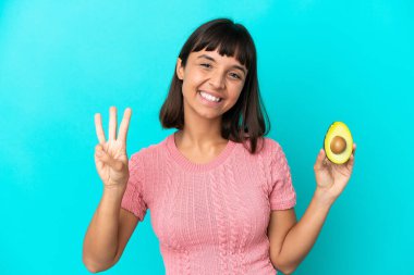 Young mixed race woman holding an avocado isolated on blue background happy and counting three with fingers