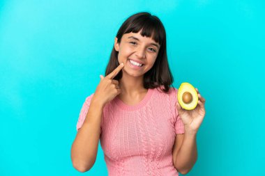 Young mixed race woman holding an avocado isolated on blue background giving a thumbs up gesture