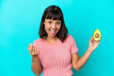Young mixed race woman holding an avocado isolated on blue background making money gesture