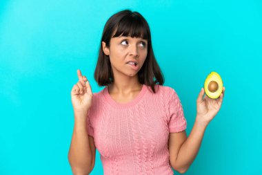 Young mixed race woman holding an avocado isolated on blue background with fingers crossing and wishing the best