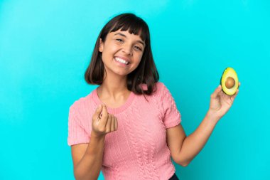 Young mixed race woman holding an avocado isolated on blue background making money gesture
