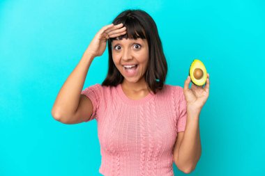 Young mixed race woman holding an avocado isolated on blue background doing surprise gesture while looking to the side