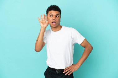 Genç Afrikalı Amerikalı bir adam mavi arka planda izole edilmiş. Kulağına elini sürerek bir şeyler dinliyor.