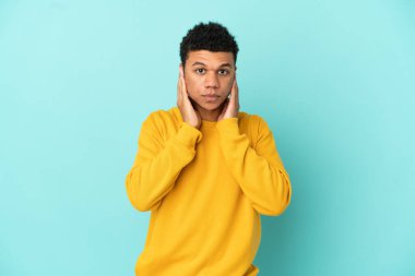 Genç Afrikalı Amerikalı adam mavi arka planda izole edilmiş hayal kırıklığına uğramış ve kulaklarını kapatmış.