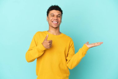 Young African American man isolated on blue background holding copyspace imaginary on the palm to insert an ad and with thumbs up