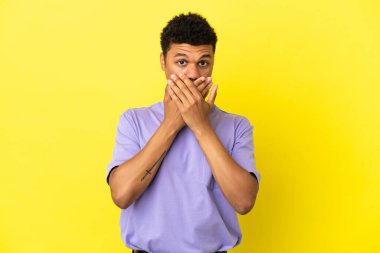 Genç Afrikalı Amerikalı adam sarı arka planda izole edilmiş ağzı elleriyle kapatıyor.