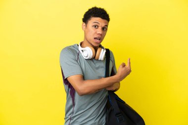Sarı arka planda izole edilmiş spor çantalı genç Afrikalı Amerikalı adam şaşırmış ve işaret ediyor.