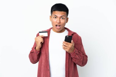 Genç Ekvadorlu adam beyaz arka planda izole edilmiş cep telefonuyla alışveriş yapıyor ve elinde şaşırmış bir ifadeyle bir kredi kartı tutuyor.