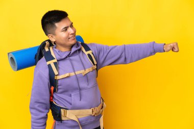 Sarı arka planda izole edilmiş büyük bir sırt çantası olan dağcı genç adam baş parmak işareti yapıyor.