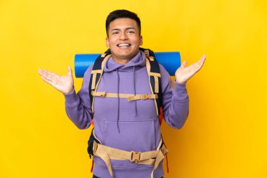 Sarı arka planda izole edilmiş büyük sırt çantalı genç dağcı, şok olmuş yüz ifadesi var.