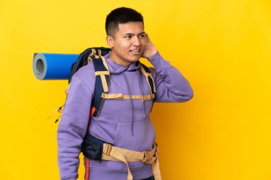 Sarı arka planda izole edilmiş büyük sırt çantalı genç dağcı elini kulağına koyarak bir şeyler dinliyor.