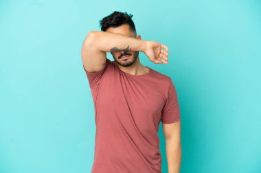 Genç beyaz adam mavi arka planda izole edildi. Gözlerini elleriyle kapadı.