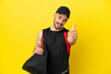 Spor çantalı genç beyaz adam sarı arka planda izole edildi. Başparmakları havada çünkü iyi bir şey oldu.