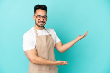 Restoran garsonu Kafkasyalı adam mavi arka planda izole edilmiş. Davet için ellerini yana uzatıyor.