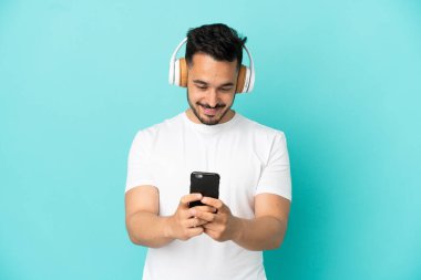 Kafkasyalı genç adam mavi arka planda izole edilmiş müzik dinliyor ve cep telefonu arıyor.