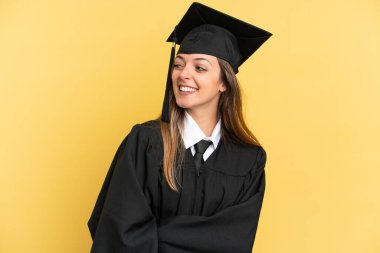 Sarı arka planda izole edilmiş genç üniversite mezunu. Yan tarafa bakıyor ve gülümsüyor.
