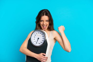 Mavi arka planda, tartı makinesiyle izole edilmiş beyaz bir kadın zafer jesti yapıyor.