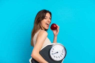 Mavi arka planda, tartı makinesi ve elmasıyla izole edilmiş genç beyaz bir kadın.