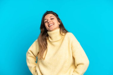 Mavi arka planda izole edilmiş genç beyaz kadın kolları kalçasında ve gülümsüyor.