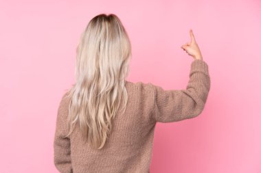 İzole edilmiş pembe arka planda süveter giyen genç sarışın kız işaret parmağıyla işaret ediyor.
