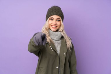 Kışlık şapkalı, mor arka plandaki genç sarışın kız kendine güvenen bir ifadeyle seni işaret ediyor.