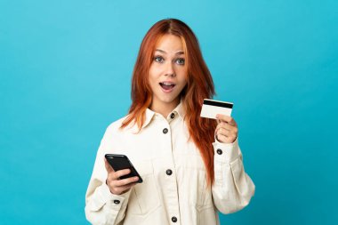 Genç Rus kız mavi arka planda izole edilmiş cep telefonuyla alışveriş yapıyor ve elinde şaşırmış bir ifadeyle bir kredi kartı tutuyor.