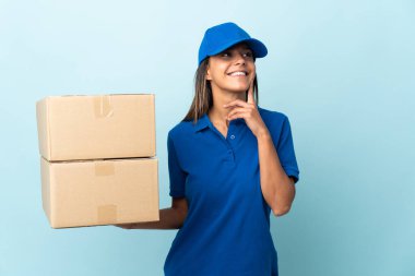 Genç teslimatçı kadın mavi arka planda izole edilmiş bir şekilde bakarken bir fikir düşünüyor.
