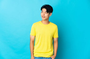 Genç Çinli adam mavi arka planda izole edilmiş, yan tarafa bakıyor ve gülümsüyor.