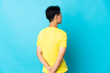 Young Chinese man isolated on blue background in back position and looking back