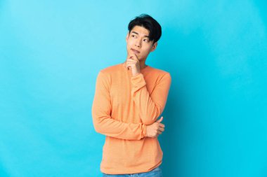 Genç Çinli bir adam mavi arka planda izole edilmiş. Şüpheleri ve düşünceleri var.