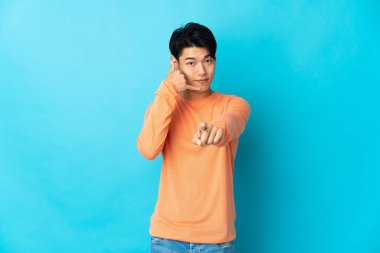 Young Chinese man isolated on blue background making phone gesture and pointing front