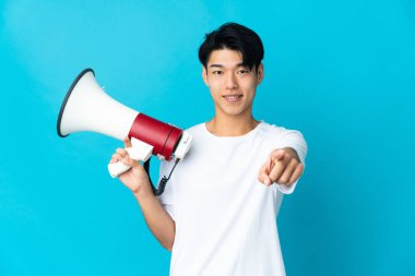 Young Chinese man isolated on blue background holding a megaphone and smiling while pointing to the front