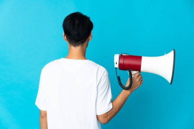 Young Chinese man isolated on blue background holding a megaphone and in back position