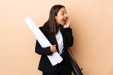 Young architect woman holding blueprints over isolated background shouting with mouth wide open to the side
