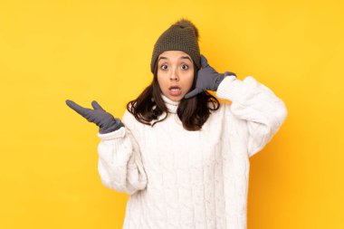 Young woman with winter hat over isolated yellow background making phone gesture and doubting
