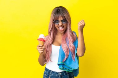 Young mixed race woman with pink hair holding ice cream isolated on yellow background celebrating a victory