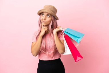 Young mixed race woman with shopping bag isolated on pink background having doubts while looking up