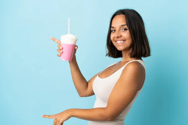 側に指を指して 製品を提示青い背景に隔離されたイチゴミルクセーキを持つ若い女性 — ストック写真