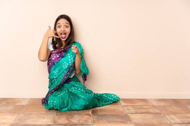 Genç Hintli bir kadın yerde oturuyor, telefon hareketleriyle ön tarafı işaret ediyor.